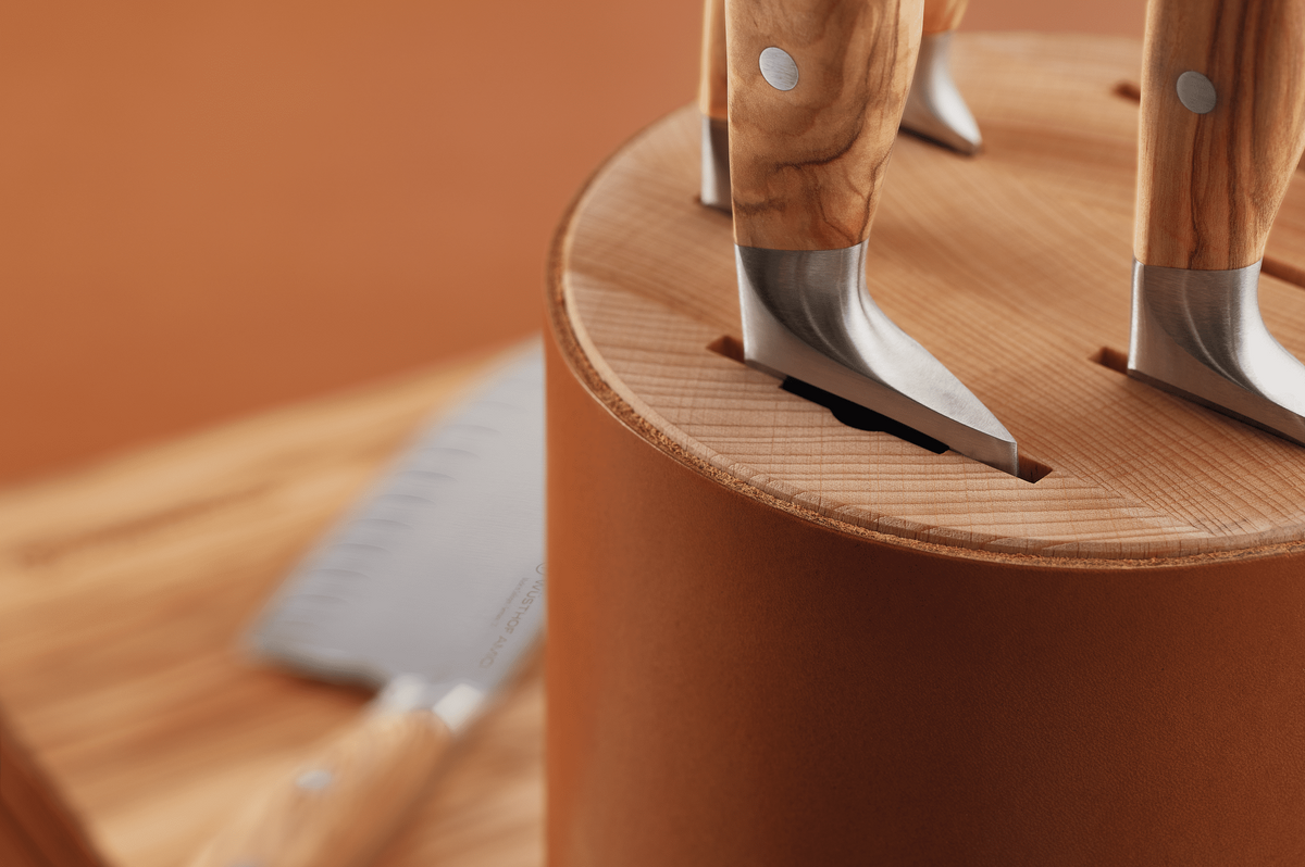 Close-up of WÜSTHOF AMICI knives standing in the round tan block, with a knife on a wooden board behind