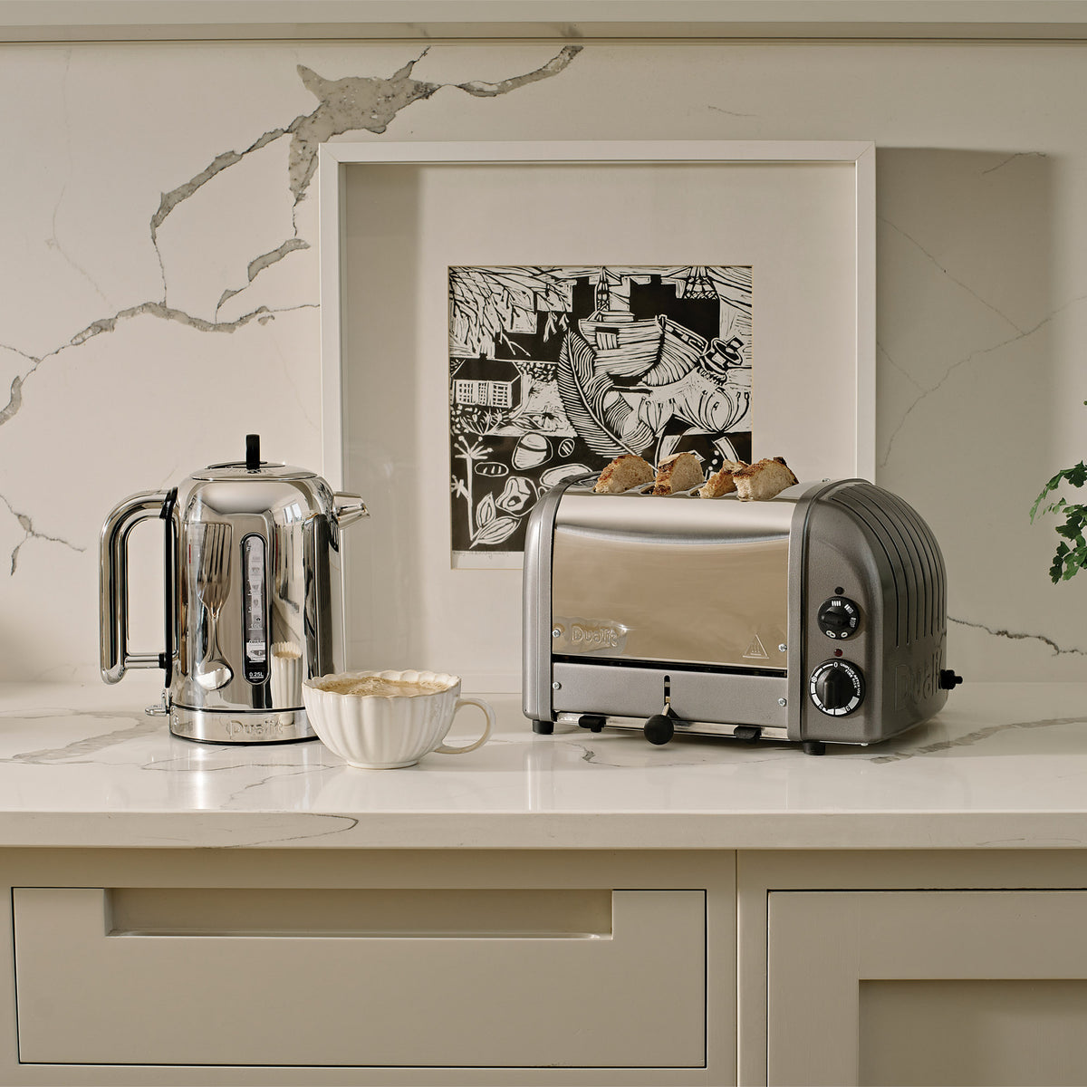 Dualit Classic 4-slice toaster in metallic charcoal with toast beside a Dualit Classic kettle on a marble counter, with a framed print