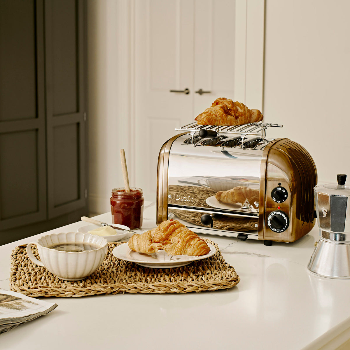 Dualit Classic 4-slice toaster in copper with a warming rack of croissants, a moka pot and jam on a kitchen island