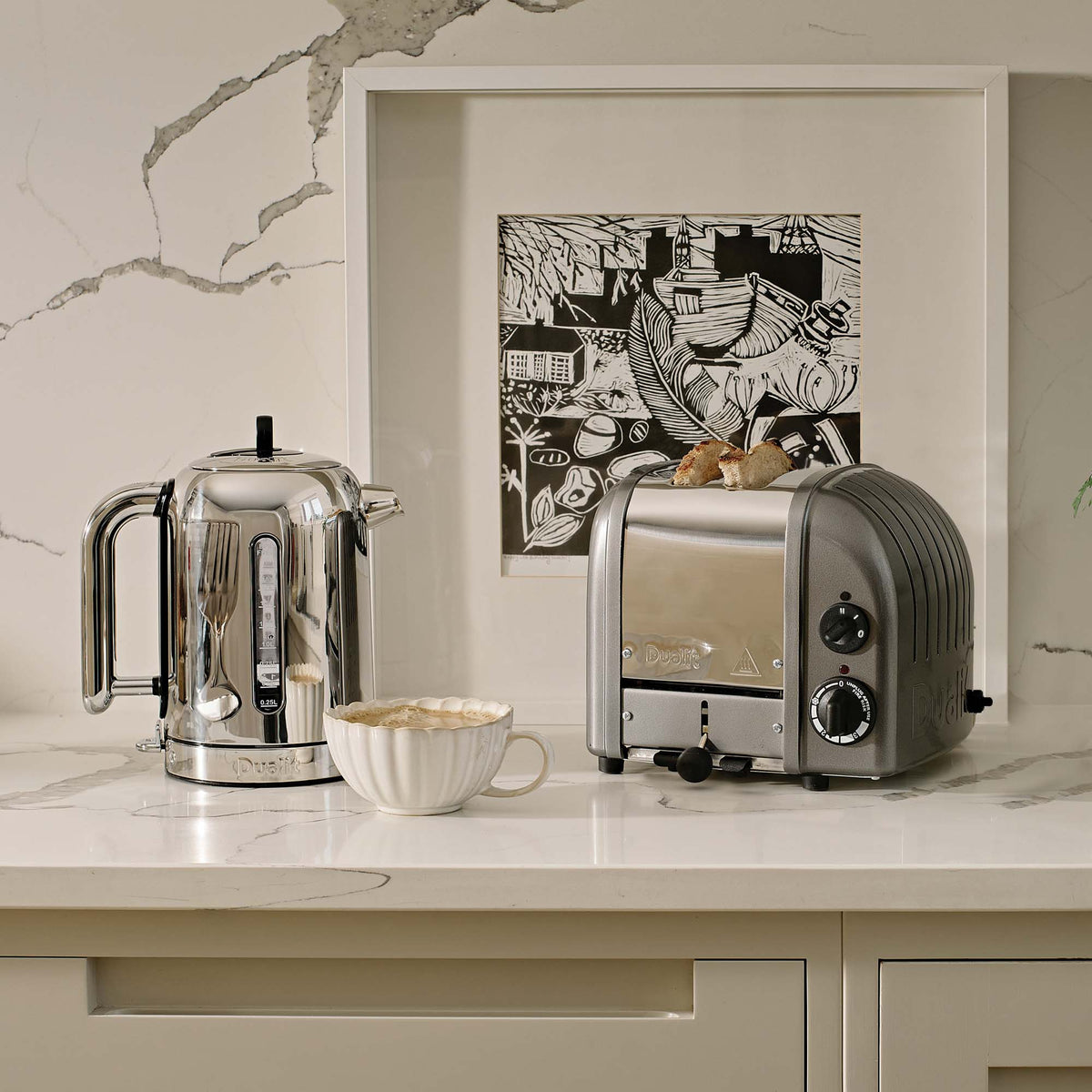 Dualit Classic 2-slice toaster in metallic charcoal beside a Dualit Classic kettle on a marble counter, with a framed print and a cup of coffee