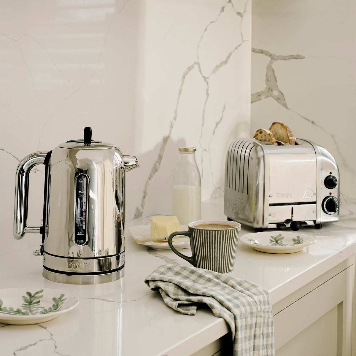 Dualit Classic kettle and 2-slice toaster in polished chrome on a marble counter with a bottle of milk, butter and a mug of coffee