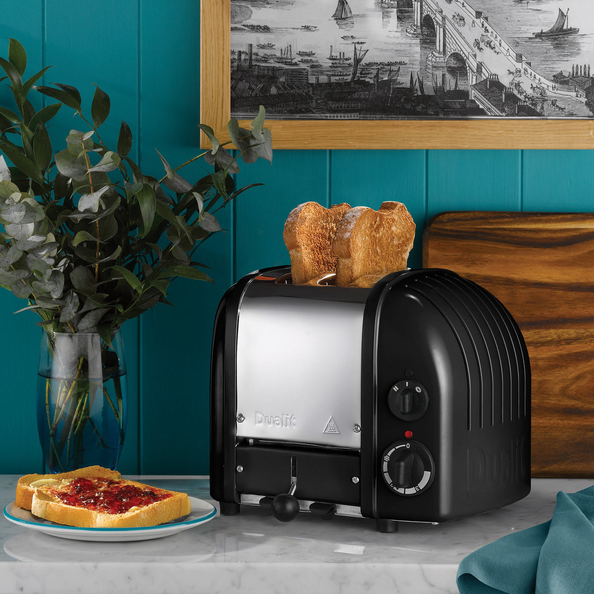 Dualit Classic 2-slice toaster in matt black with toast on a marble counter against a teal panelled wall, with jam on toast and eucalyptus