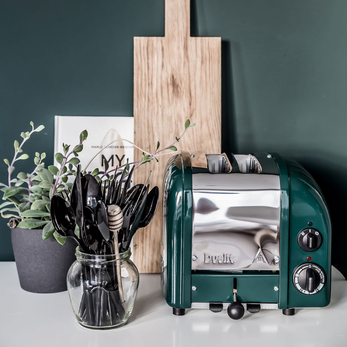Dualit Classic 2-slice toaster in evergreen beside a jar of black cutlery and a wooden board against a green wall