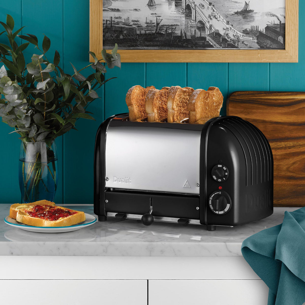 Dualit Classic 4-slice toaster in matt black with toast on a marble counter against a teal panelled wall, with jam on toast and eucalyptus