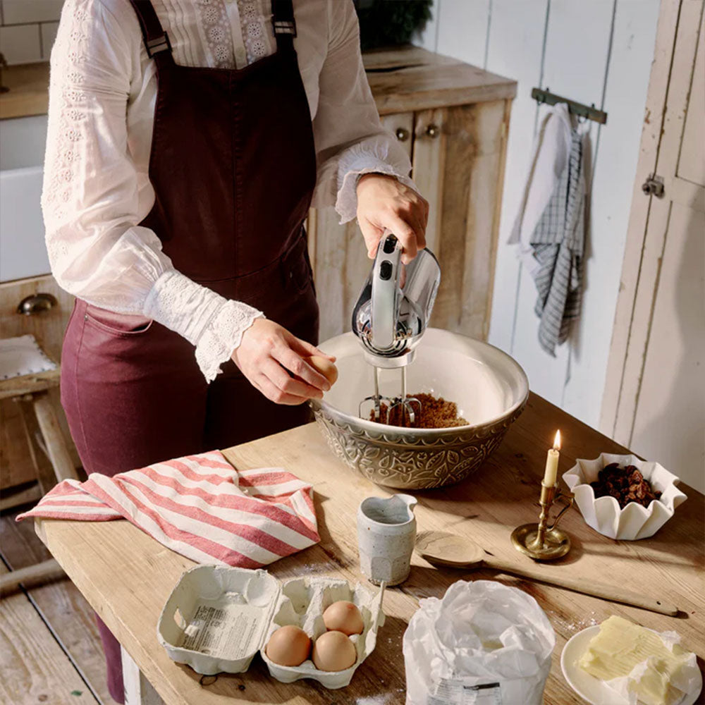 Woman mixing cake batter with the Dualit hand mixer at a rustic wooden table with eggs