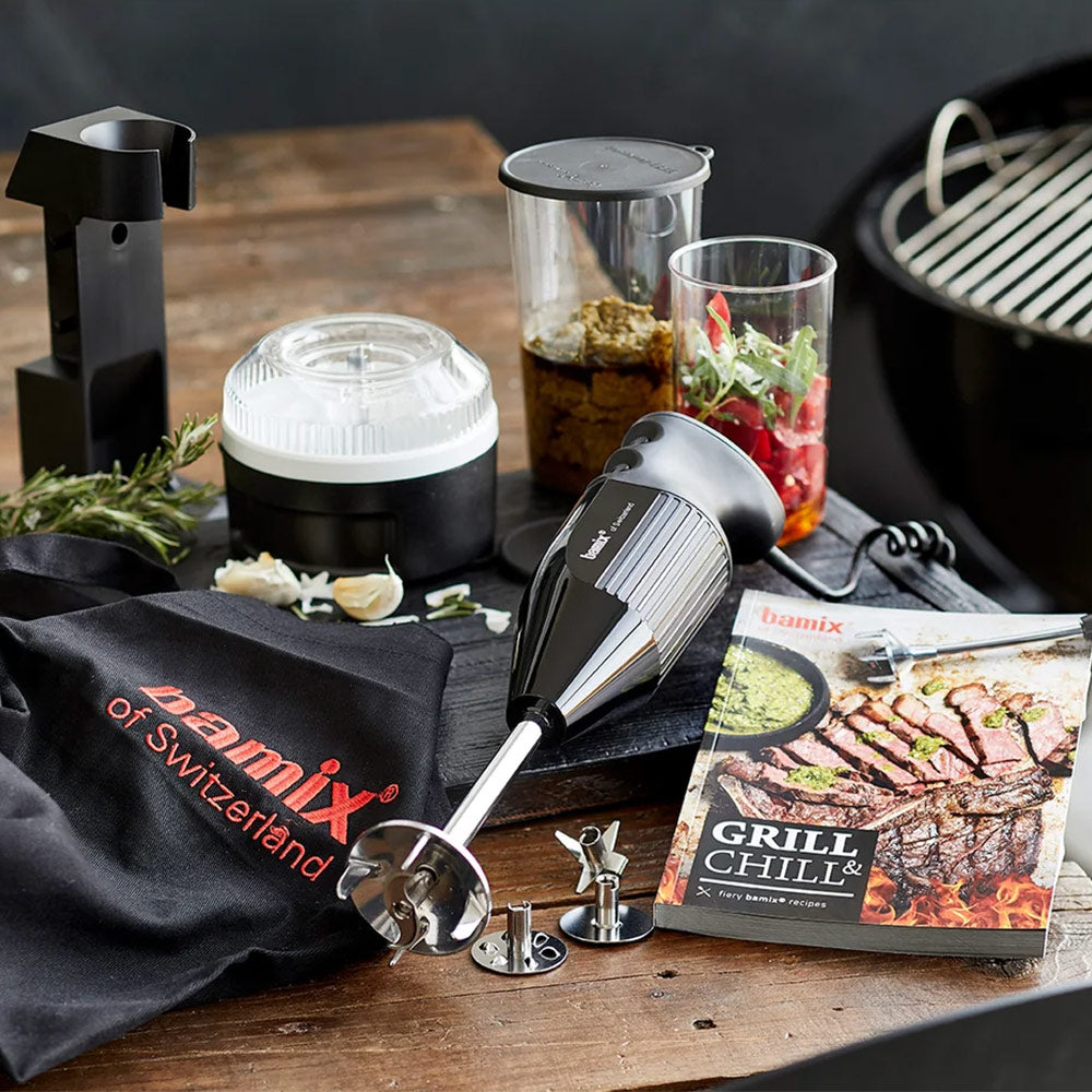 bamix BBQ hand blender with its attachments, Processor, apron and Grill & Chill book on a wooden table beside a grill