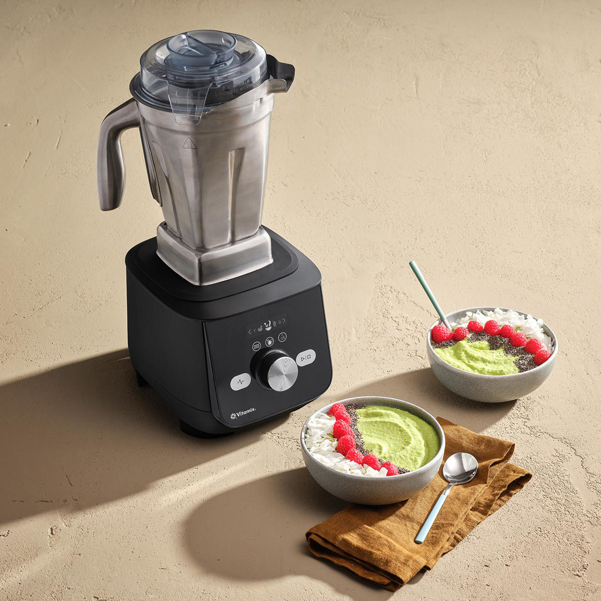 Vitamix 48 oz Stainless Steel Container on a black Vitamix blender, with green smoothie bowls topped with raspberries
