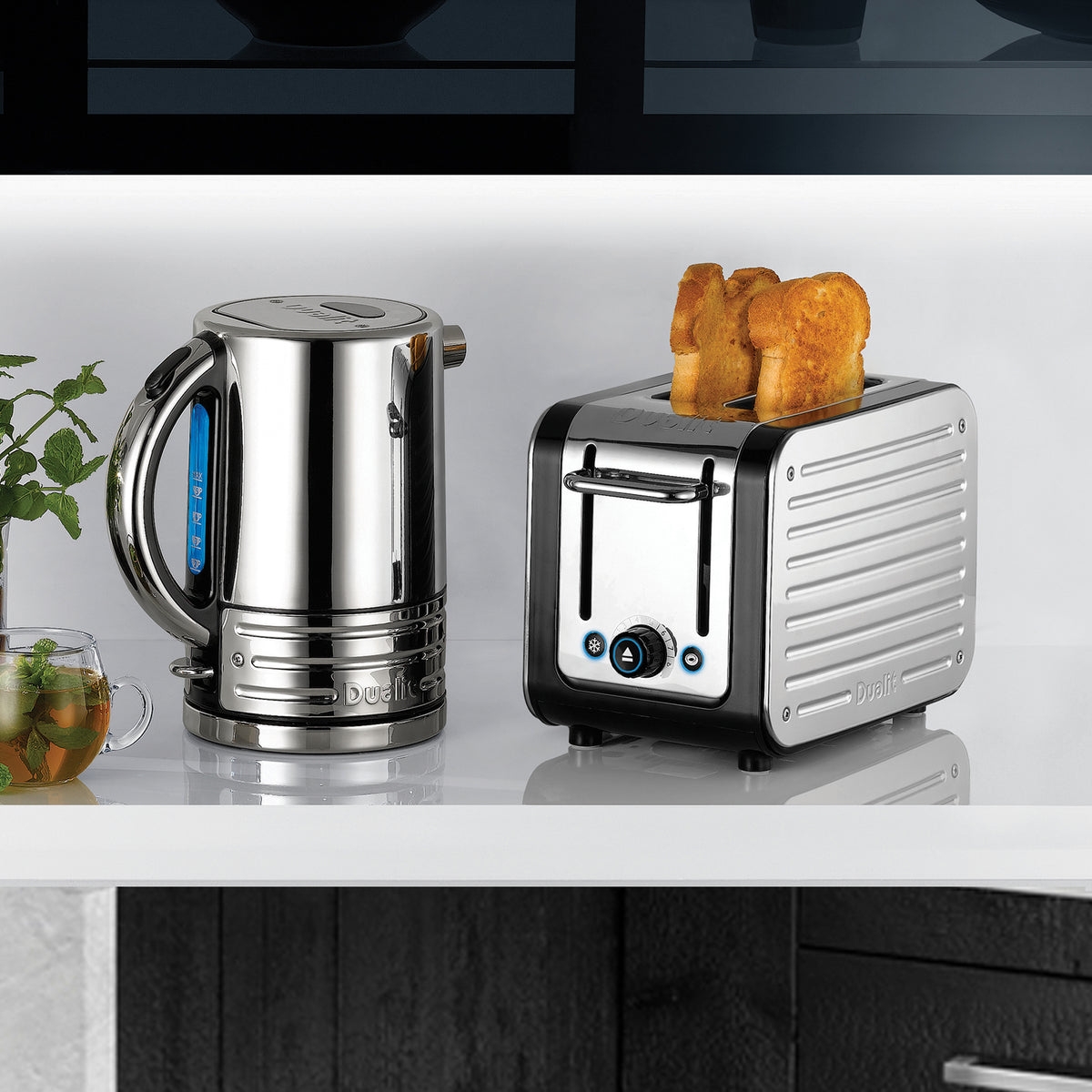 Dualit Design Series 2-slice toaster with toast beside the matching kettle and a glass of mint tea on a white shelf