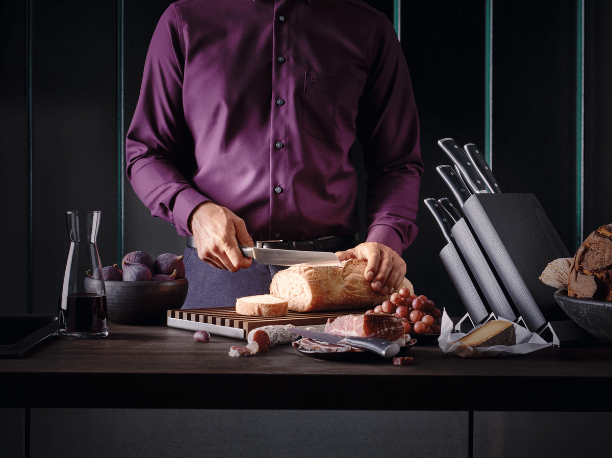 Man in a purple shirt slicing bread at a counter, with WÜSTHOF CLASSIC IKON knives in a block beside him