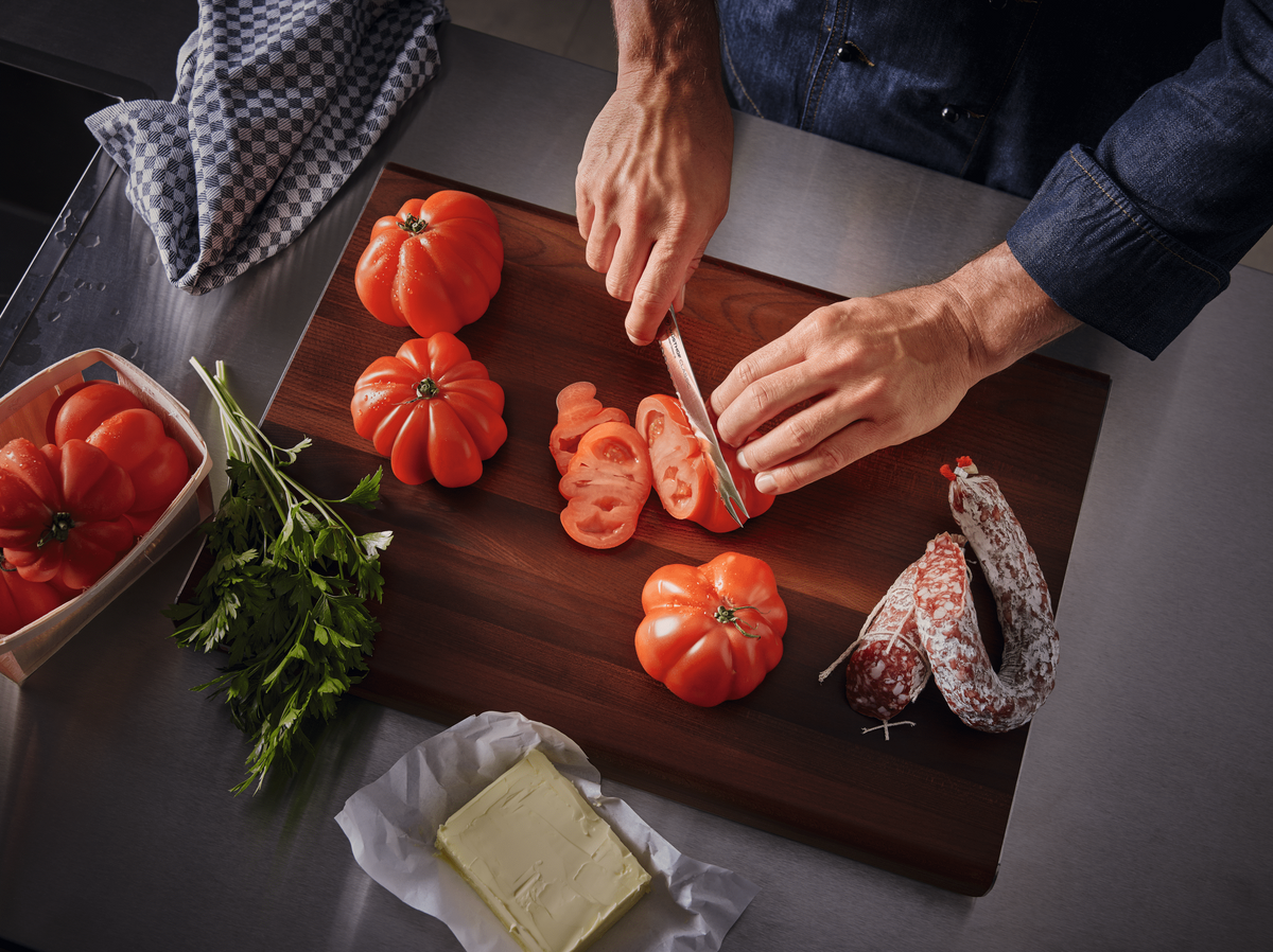 Hands slicing tomatoes with a WÜSTHOF CLASSIC knife on a dark wooden board, with parsley, salami and butter