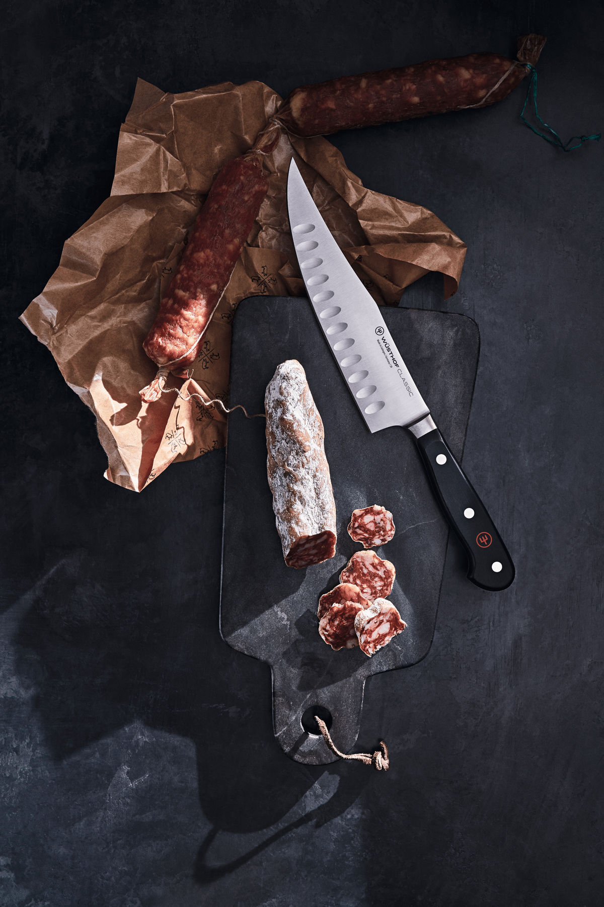 WÜSTHOF CLASSIC 7" Hollow Edge Craftsman on a dark board with sliced salami and brown paper