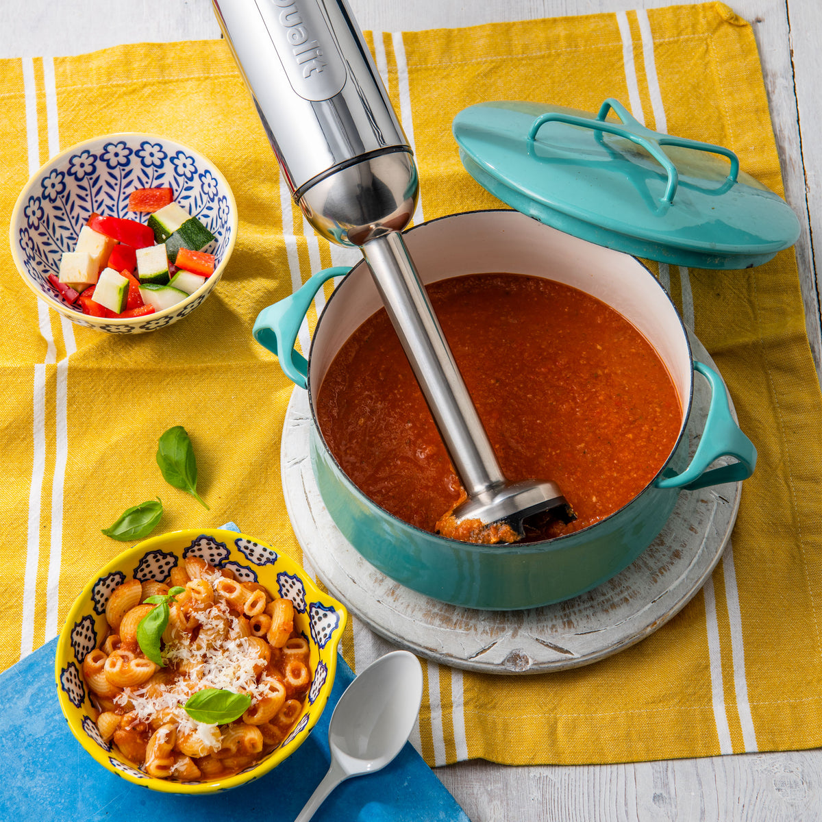 Dualit hand blender in a teal pot of tomato soup, seen from above with pasta and vegetables