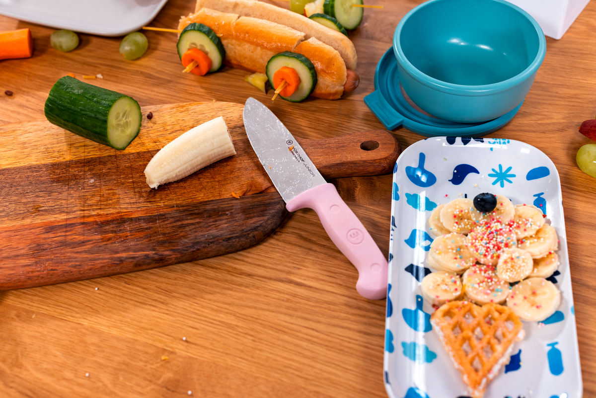 WÜSTHOF Junior 4" Chef's Knife, Pink, on a wooden board with cucumber and a child's plate of banana and waffle